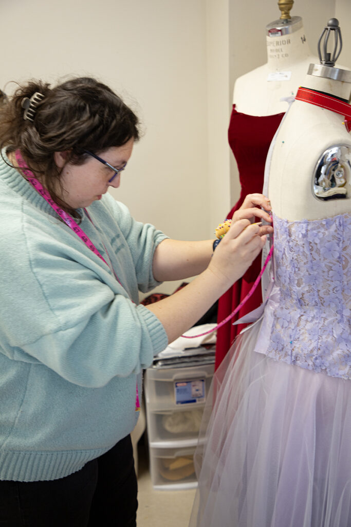 A person stands next to a mannequin that has a lavender dress on it. She holds a measuring tape against a section of the dress.