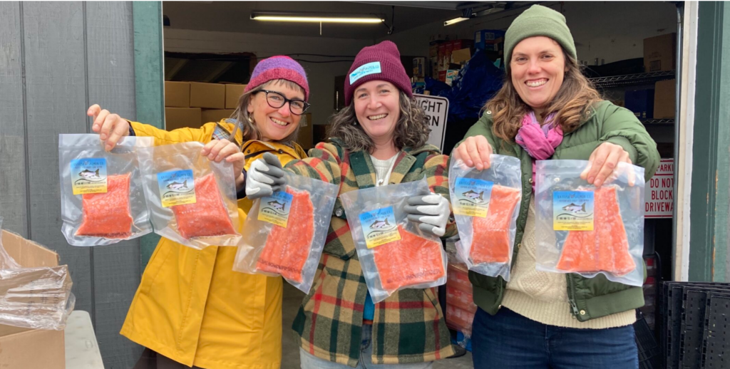 Three people, bundled in warm outfits and smiling, hold out packages of salmon toward to the camera.