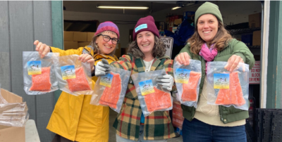 Three people, bundled in warm outfits and smiling, hold out packages of salmon toward to the camera.