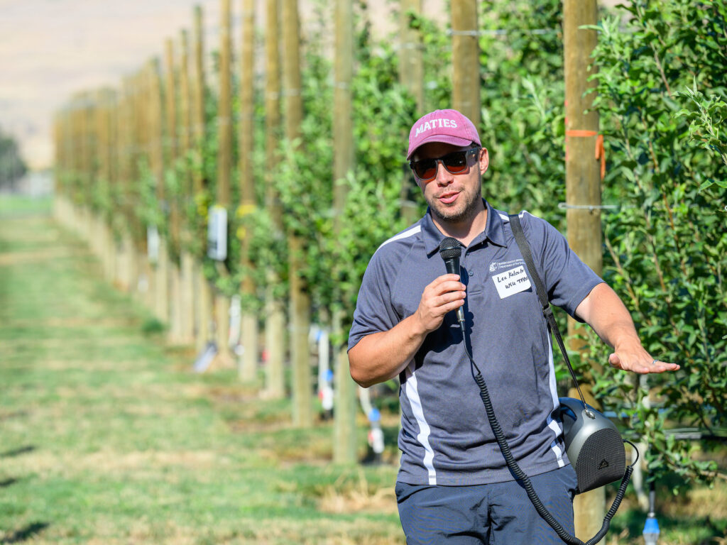 Kalcsits speaks at Wenatchee research station orchard, holding microphone.
