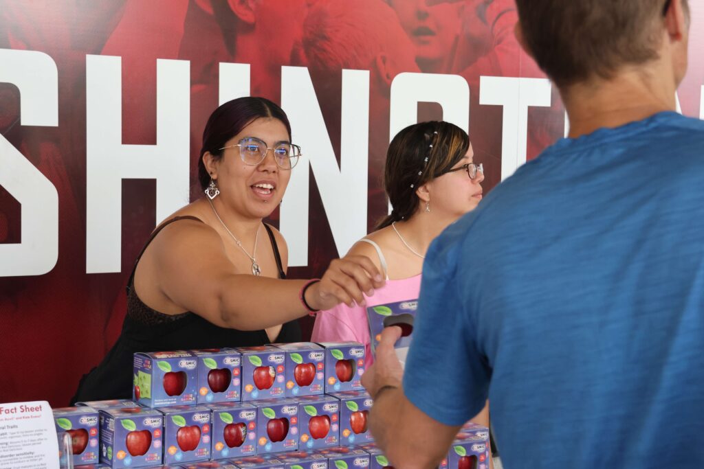 Leisly, left, with her sister facing to the right, hands a boxed apple to person visiting a table display of apples/
