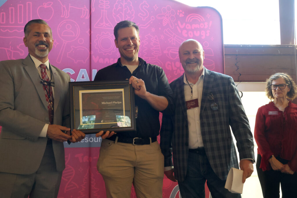 Award recipient Michael Phelps, center, holds award certificate on stage in front of WSU-themed backdrop. CAHNRS Dean Raj Khosla stands at left, helping hold the certificate. Flanking Phelps are Gordon Murdoch, with Judy Hopkins to the back of the stage at right.