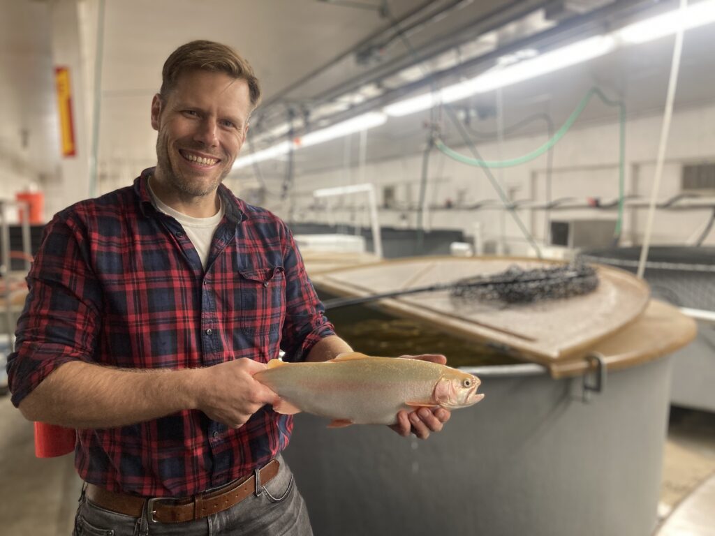 Phelps, holding a large pink-colored trout in a long room lined with pond-like tanks, conduits, and lights. A fish net rests on a nearby tank.