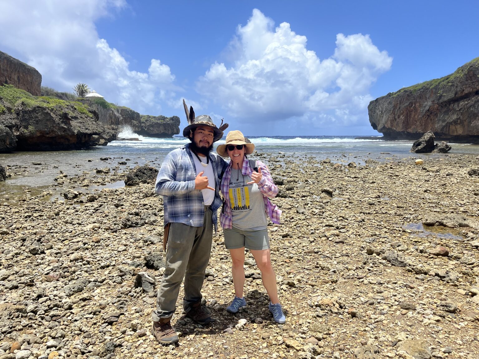 Two people pose on a rocky beach with the ocean behind them. They're both giving the “hang loose” hand sign.