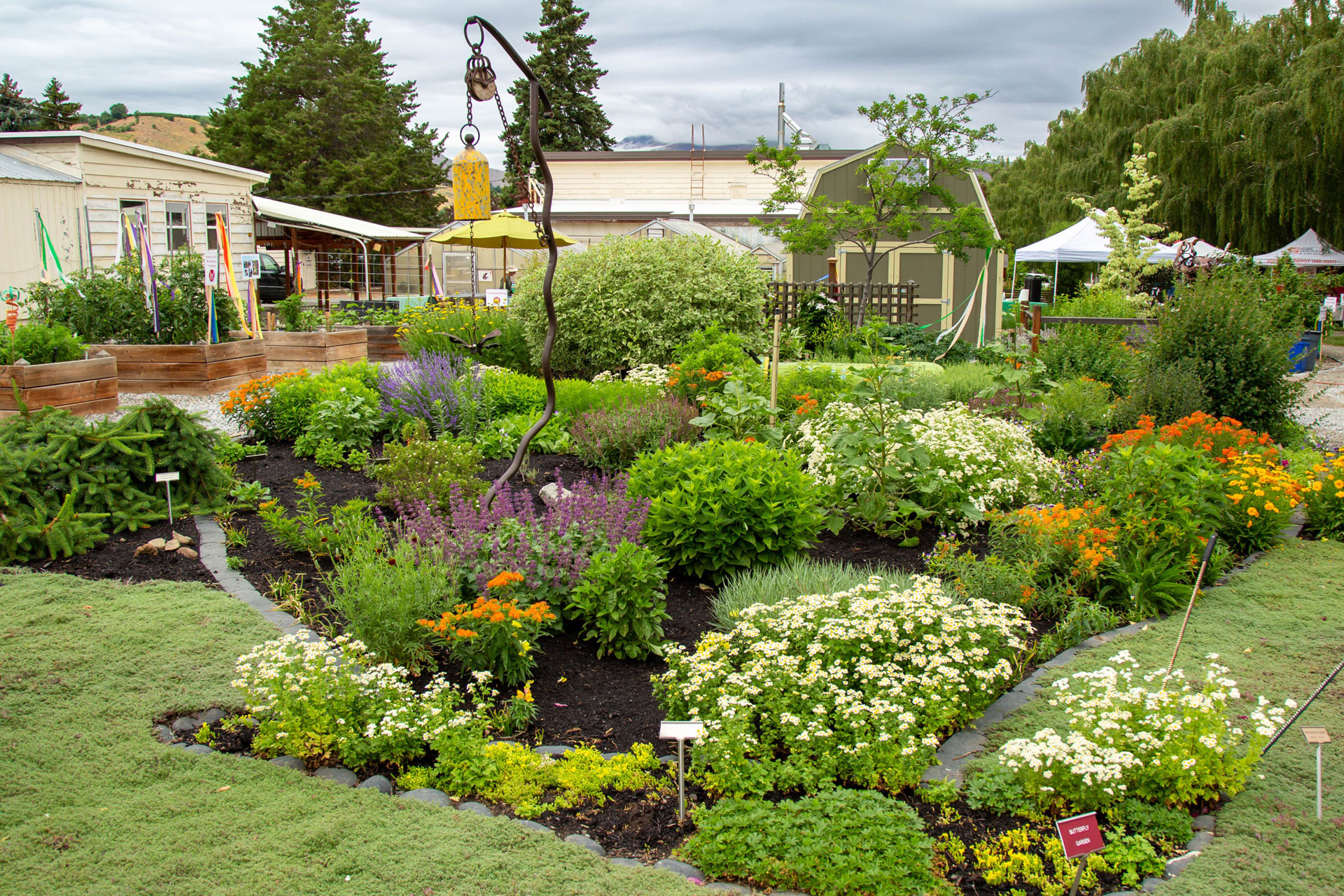  Landscaped area during spring bloom in the foreground. Buildings, garden shed, and pop-up canopies in the background.