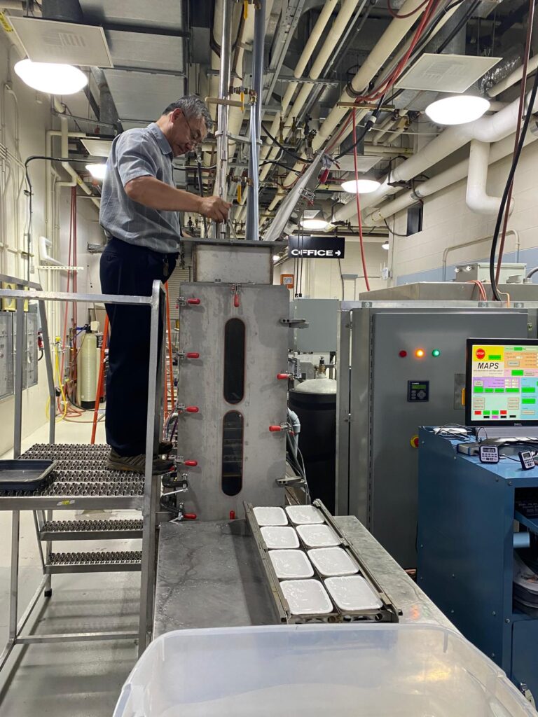 A large piece of metal equipment is behind a large tray that holds 8 small white plastic trays.