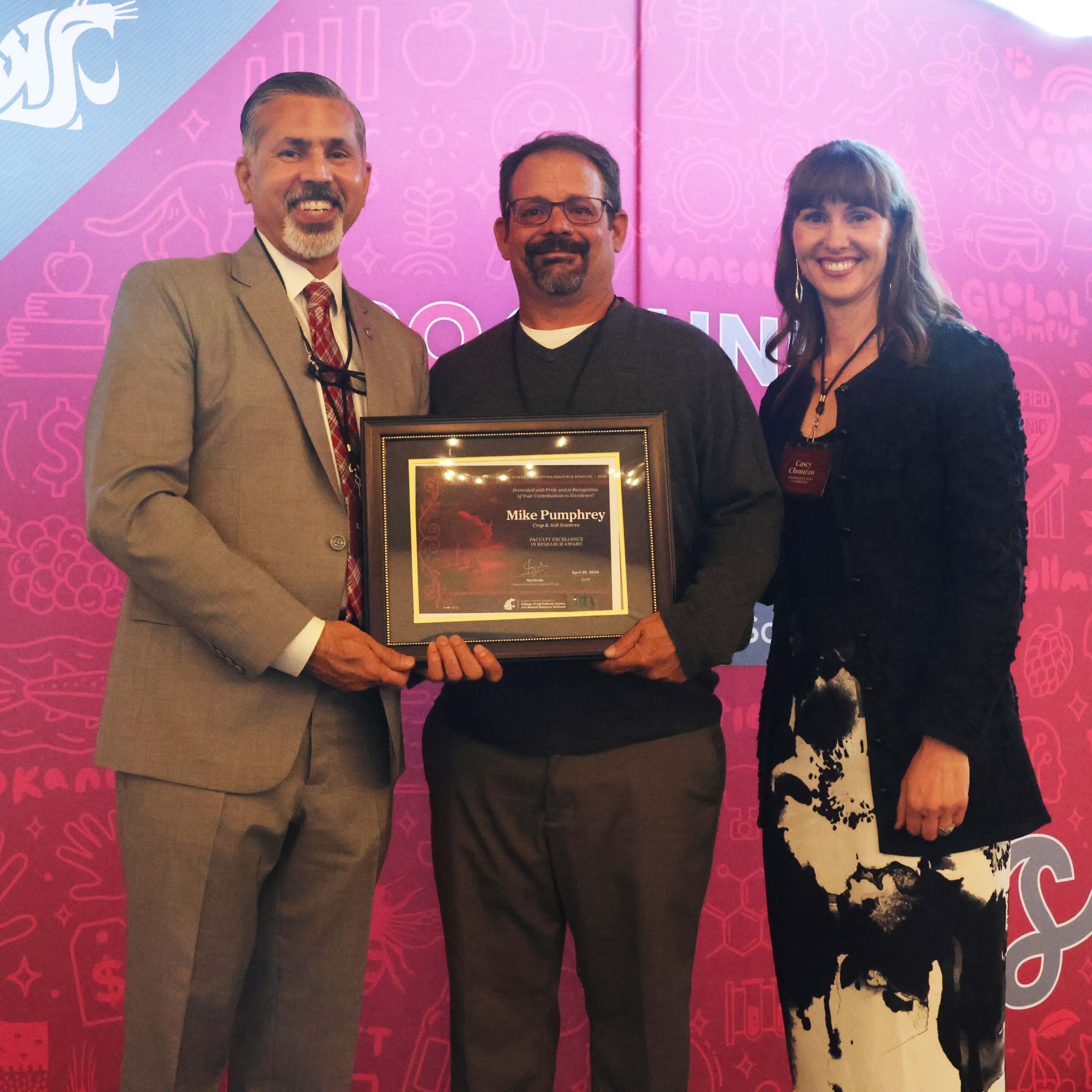 Award recipient Michael Pumphrey, center, holds award certificate on stage in front of WSU-themed backdrop. CAHNRS Dean Raj Khosla stands at left, helping hold the certificate. On the right is Casey Chumrau.