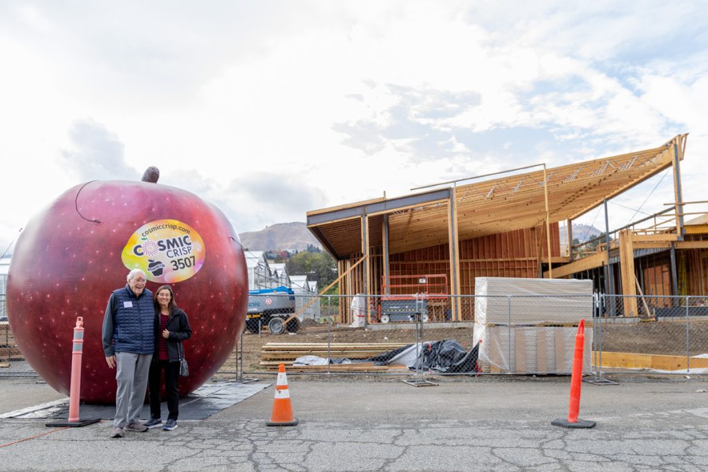 The Hambeltons stand near a large inflatable red apple. In the background is a building under construction with exposed structure and a fence.