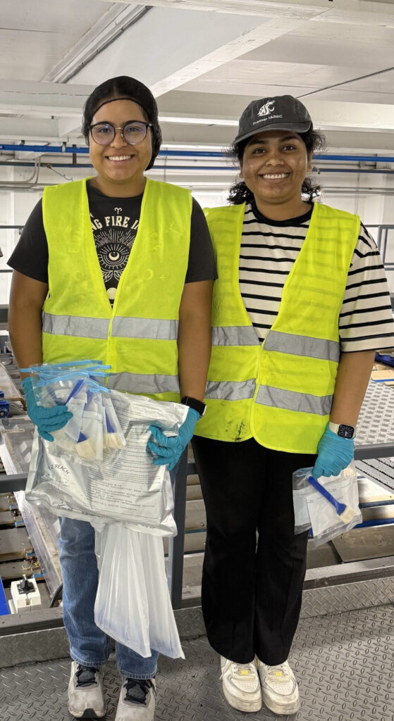Ruiz and Medikonda, smiling and facing the camera, wearing head coverings and fluorescent vests, holding plastic kits containing pouches and small brushes.