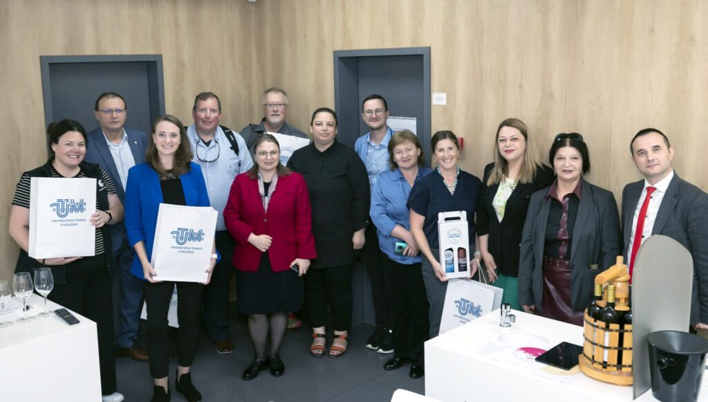 A group of 13 people stands together. Three people hold bags that say "UTM" on them. One person also holds a box of two bottles of wine. There is a table with a few bottles of wine on it, and a table with empty wine glases on it. Behind the group are two doors and a wood paneled wall.
