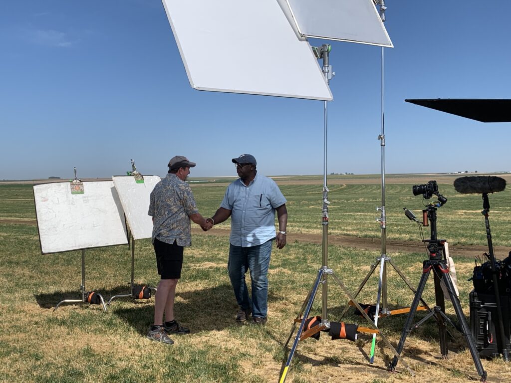 2 people shake hands in a field. They are surrounded by television equipment.