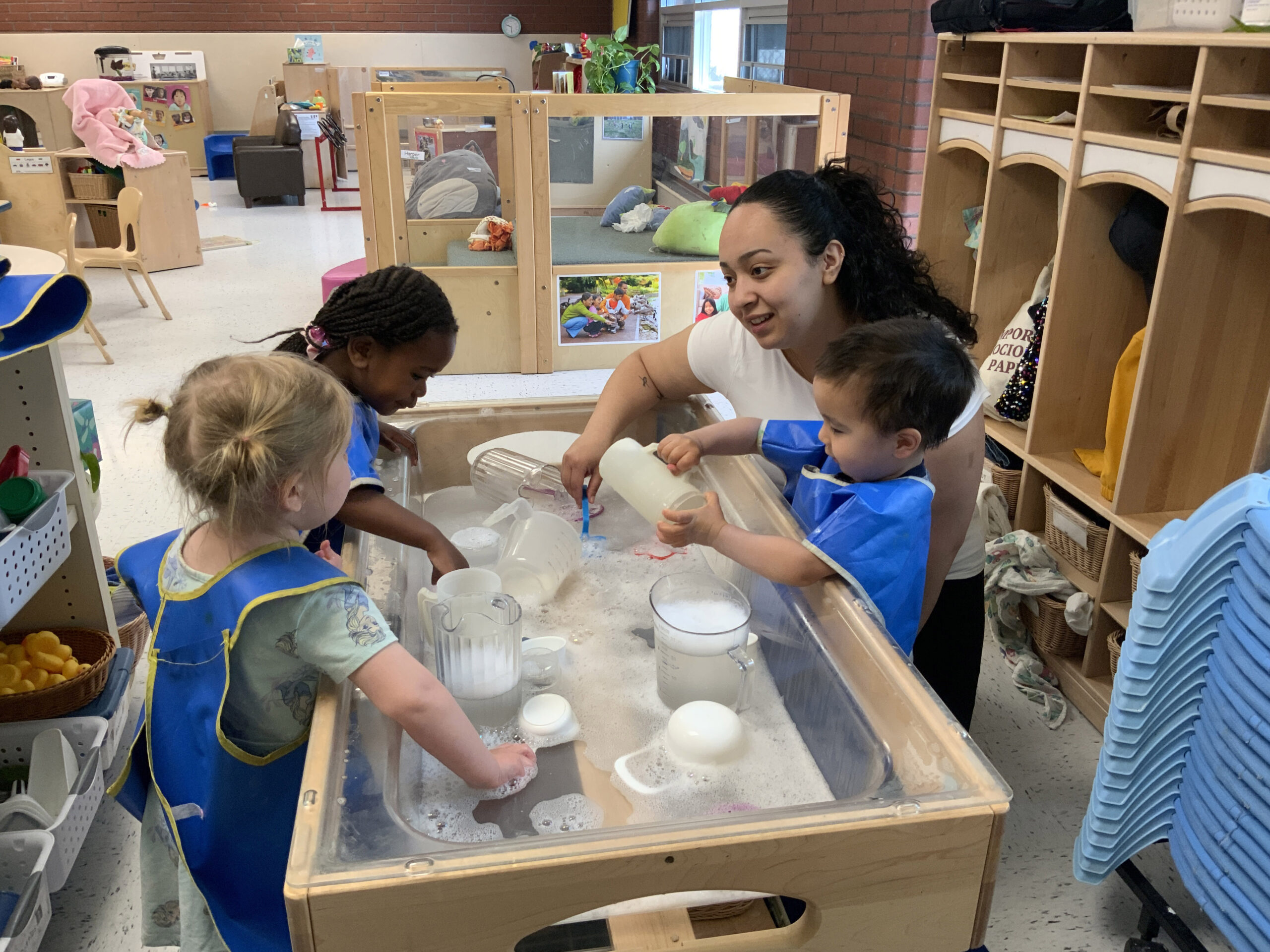 An adult is surrounded by 3 small children as they all play with bubbles at a small table. The children are all wearing smocks.