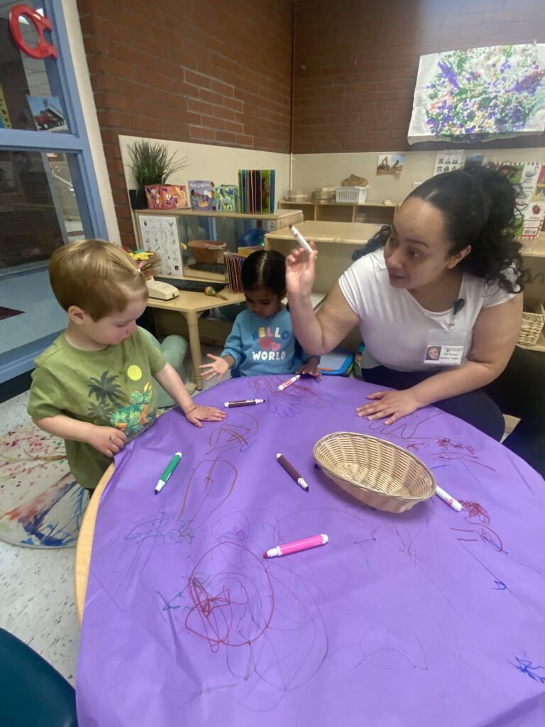 An adult sits with 2 small children at a tiny table that is covered with a large paper. The paper has many squiggles and drawings on it, including a few hands.