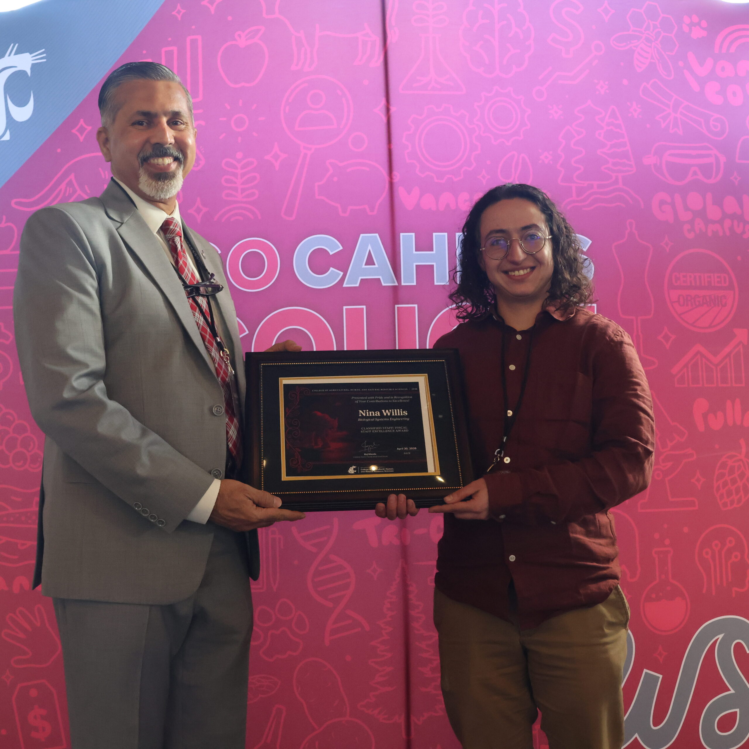 Award recipient Nina Willis, right, holds award certificate on stage in front of WSU-themed backdrop. CAHNRS Dean Raj Khosla stands at left, helping hold the certificate.