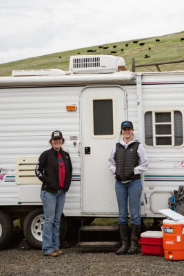 2 people stand in front of a small white camper. Cows are visible on the hill behind them.