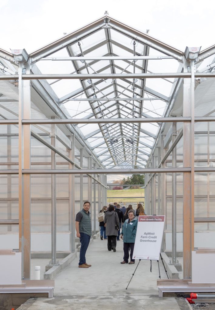 Kalcsits and Snapp, with a tour group walking away behind them, stand inside a greenhouse hallway under construction. A sign reads Plant Growth Facility AgWest Farm Credit Greenhouse.