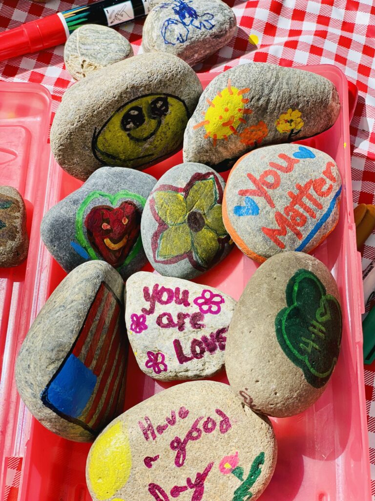 Painted rocks on a red checkered tablecloth, each featuring cheerful messages and drawings like “You Matter,” “You are Loved,” a smiling sun, a 4-H clover, and colorful hearts and flowers.
