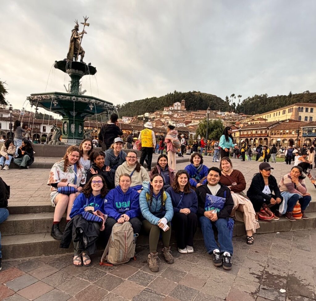 A group of roughly a dozen people sits on steps in a brick square. Behind them is a fountain and buildings with Peruvian-style architecture.