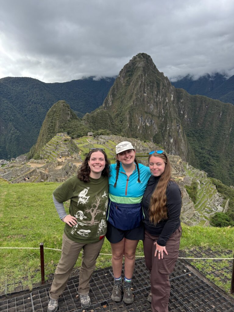 Three people stand together. Behind them are stone ruins and mountains with vegetation growing on them.
