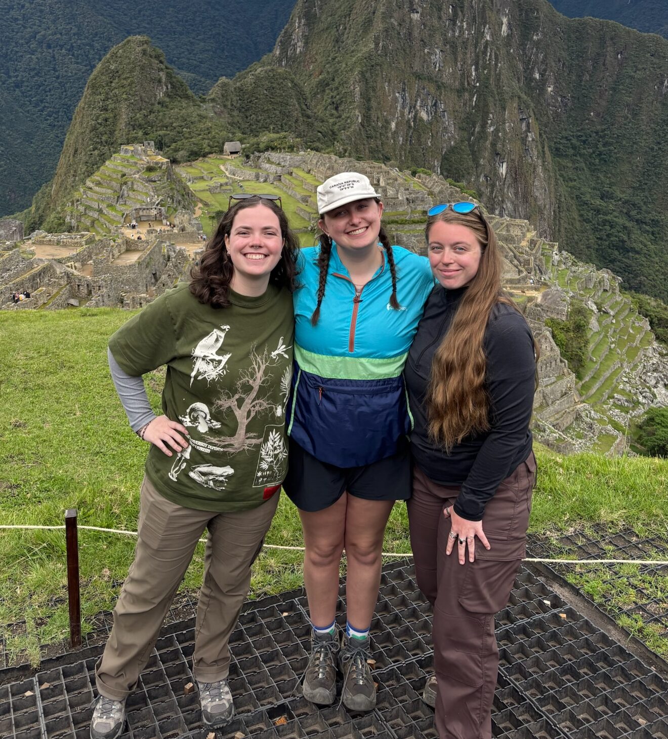 Three people stand together. Behind them are stone ruins and mountains with vegetation growing on them.