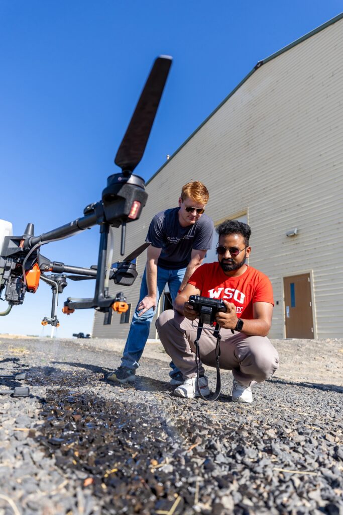 Two researchers, one crouching with a remote control, the other looking over his shoulder at the controls, stand next to a grounded aerial drone outside a large beige building.