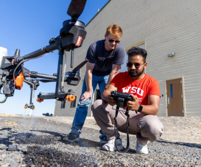 Two researchers, one crouching with a remote control, the other looking over his shoulder at the controls, stand next to a grounded aerial drone outside a large beige building.