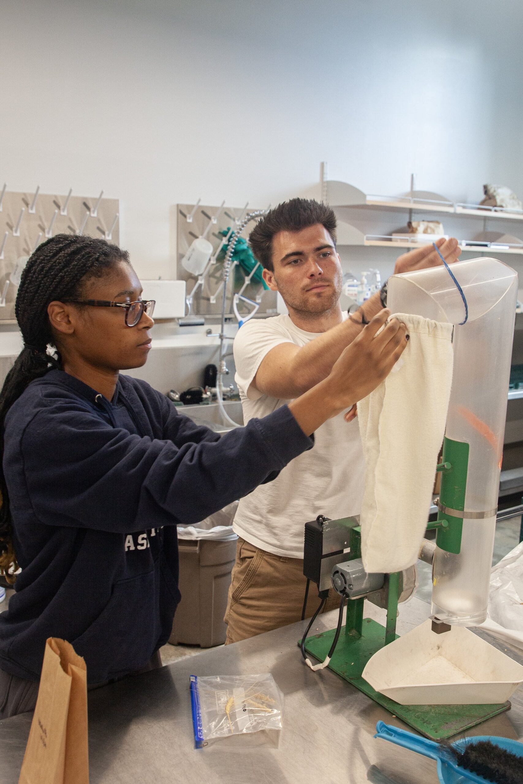 Two students working in a lab. One is holding a canvas bag up to a nozzle, while the other holds the nozzle steady.