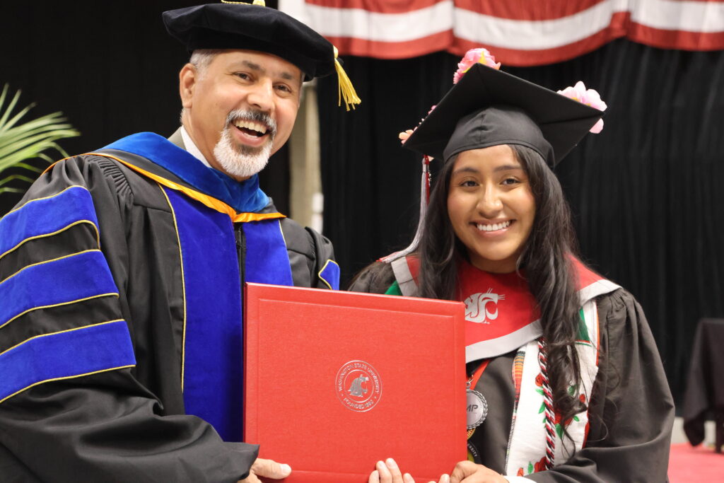 Dean Khosla, left, and Ortiz, right, in commencement gowns, smile as they together hold a closed red diploma.