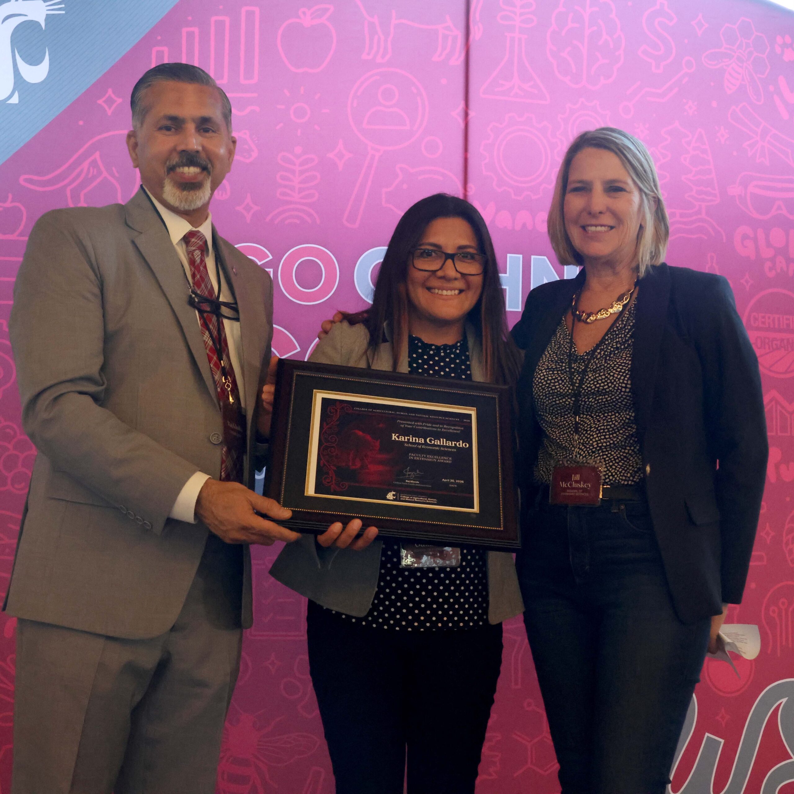 Award recipient Karina Gallardo, center, holds award certificate on stage in front of WSU-themed backdrop. CAHNRS Dean Raj Khosla stands at left, helping hold the certificate. On the right is Jill McCluskey.