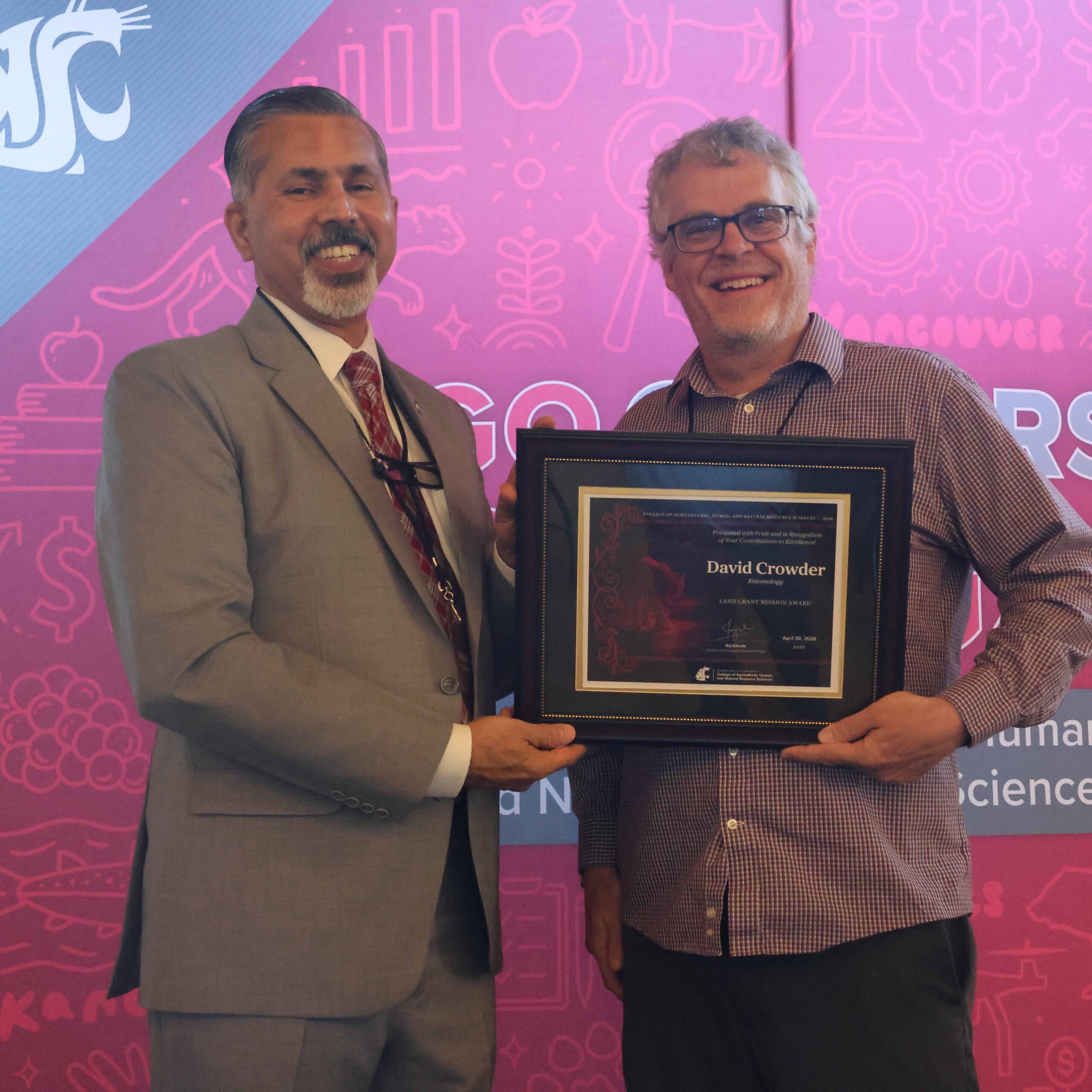 Award recipient David Crowder, right, holds award certificate on stage in front of WSU-themed backdrop. CAHNRS Dean Raj Khosla stands at left, helping hold the certificate.