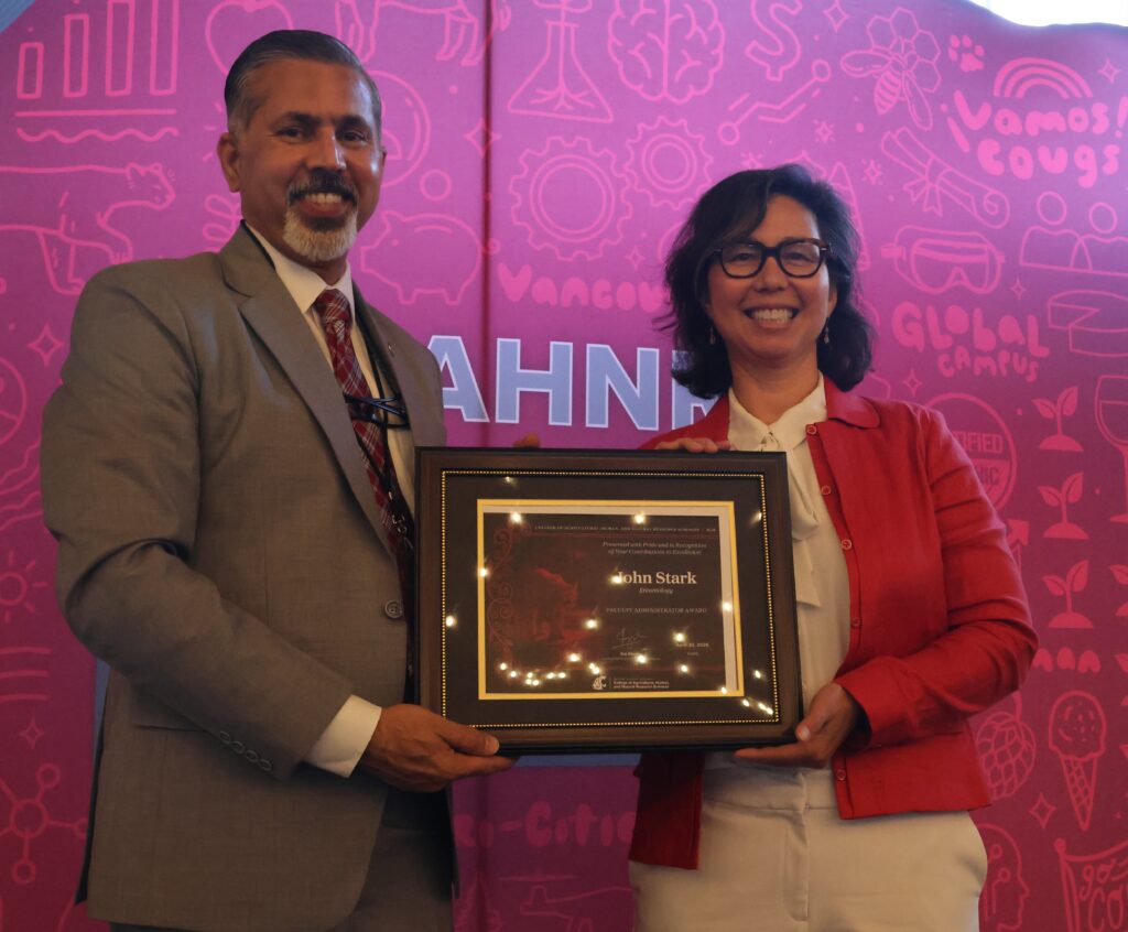 Laura Lavine, right, holds an award certificate (addressed to John Stark) on stage in front of WSU-themed backdrop. CAHNRS Dean Raj Khosla stands at left, helping hold the certificate.