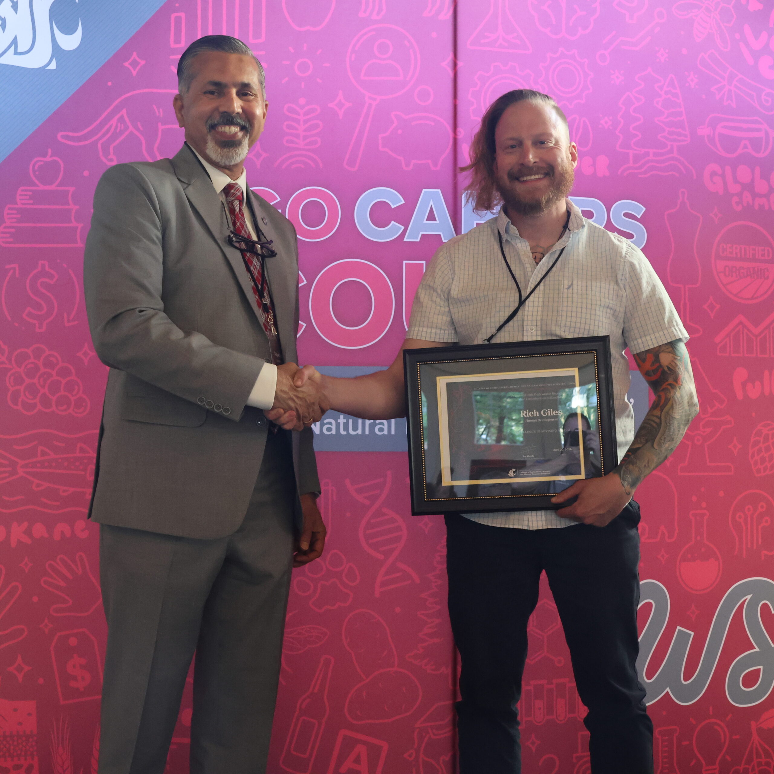 Award recipient Rich Giles, right, holds award certificate on stage in front of WSU-themed backdrop. CAHNRS Dean Raj Khosla stands at left, helping hold the certificate.