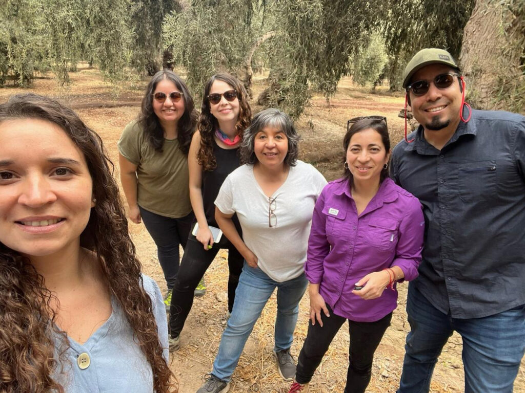 A group of five woman and a man take a selfie-style photo in a wooded, park-like outdoor area.