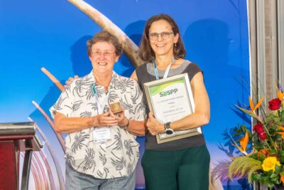 Two people stand on a stage, smiling. One holds a gold medal embedded in a wooden block, while the other holds a framed award that reads SASPP The Christaan Hendrik Persoon Medal presented to Prof Lindsay du Toit.