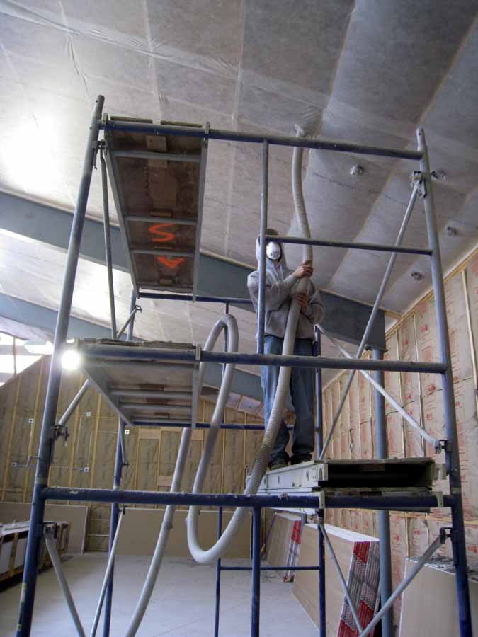 Someone on a scaffold in personal protective equipment blows in insulation via a tube into the rafters of a new-construction home.