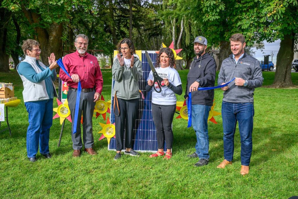 Six people stand in front of a solar panel in a park-like setting. One person holds an oversized pair of scissors, and other people hold the tail ends of a cut ribbon.