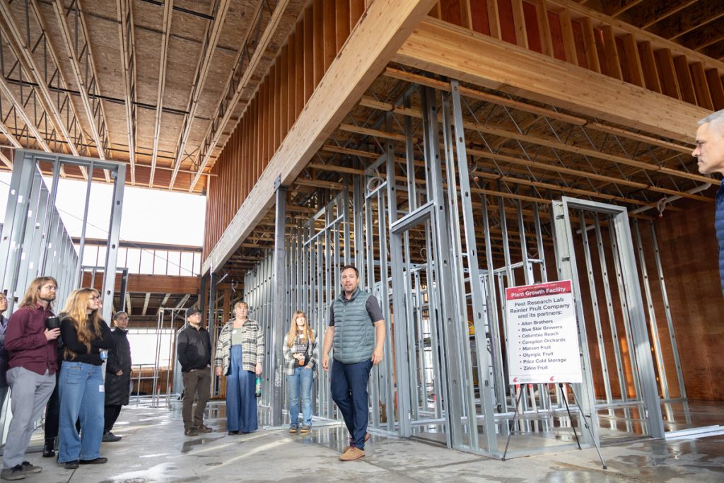 Kalcsits, in vest, stands while seven people stand and watch him, inside a large building under construction. A sign near him read Plant Growth Facility Pest Research Lab Rainier Fruit Company and its partners.