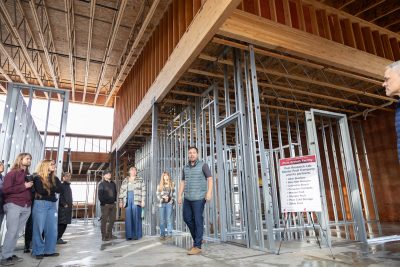 Kalcsits, in vest, stands while seven people stand and watch him, inside a large building under construction. A sign near him read Plant Growth Facility Pest Research Lab Rainier Fruit Company and its partners.
