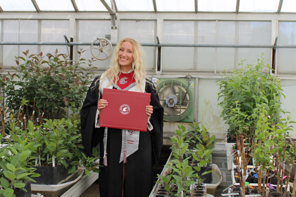 A person wearing a cap and gown holds a red leather-bound folder in a greenhouse.