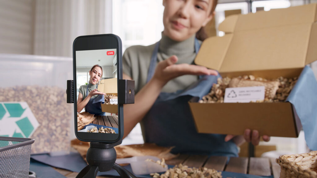 A woman holds a box with recycled packing materials while filming herself with a cell phone.