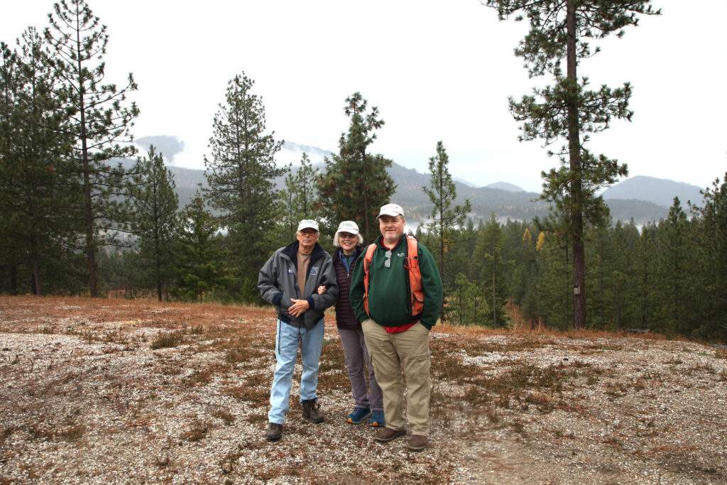 Three people stand together next to a forest. Forested hills are in the background.