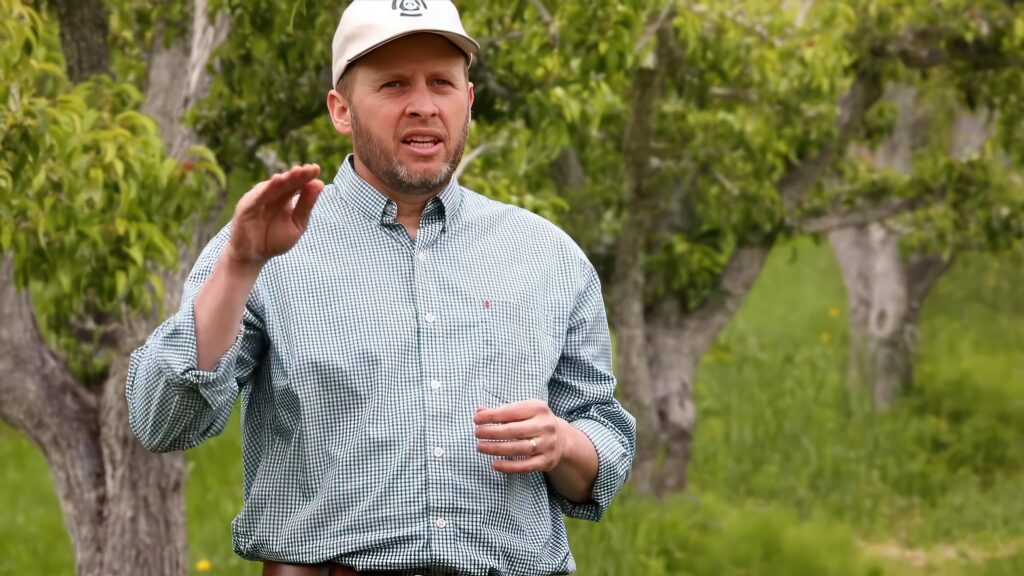 A person talking in an orchard, with trees in the background.