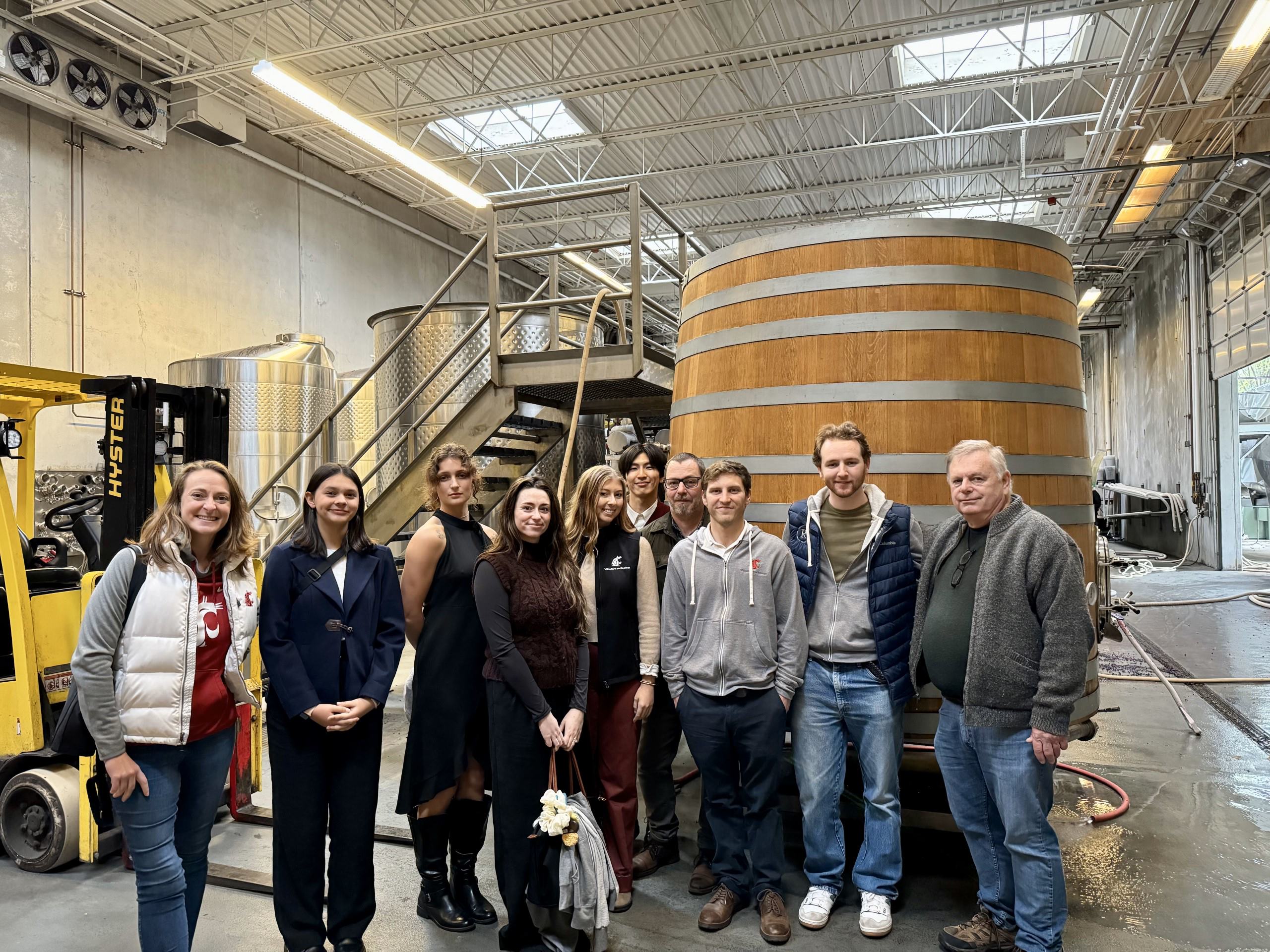 A group of 10 people stands in a winery facility, next to a large wooden barrel. Other winemaking equipment is in the background.