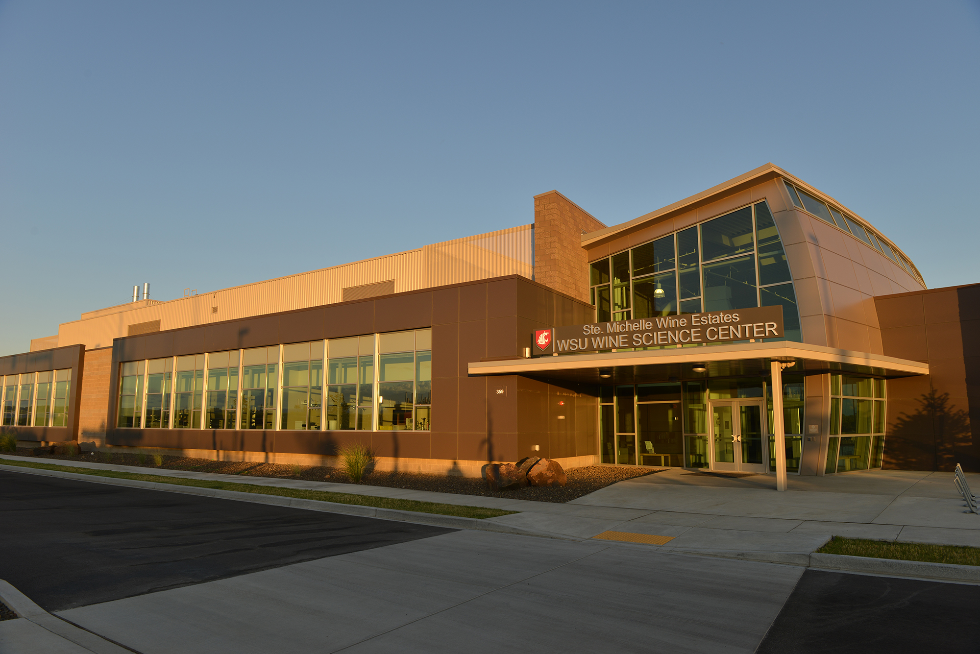 The outside of a building with sun shining on it. A sign on the building entrance includes the WSU cougar head logo and the words “Ste. Michelle Wine Estates WSU Wine Science Center.”