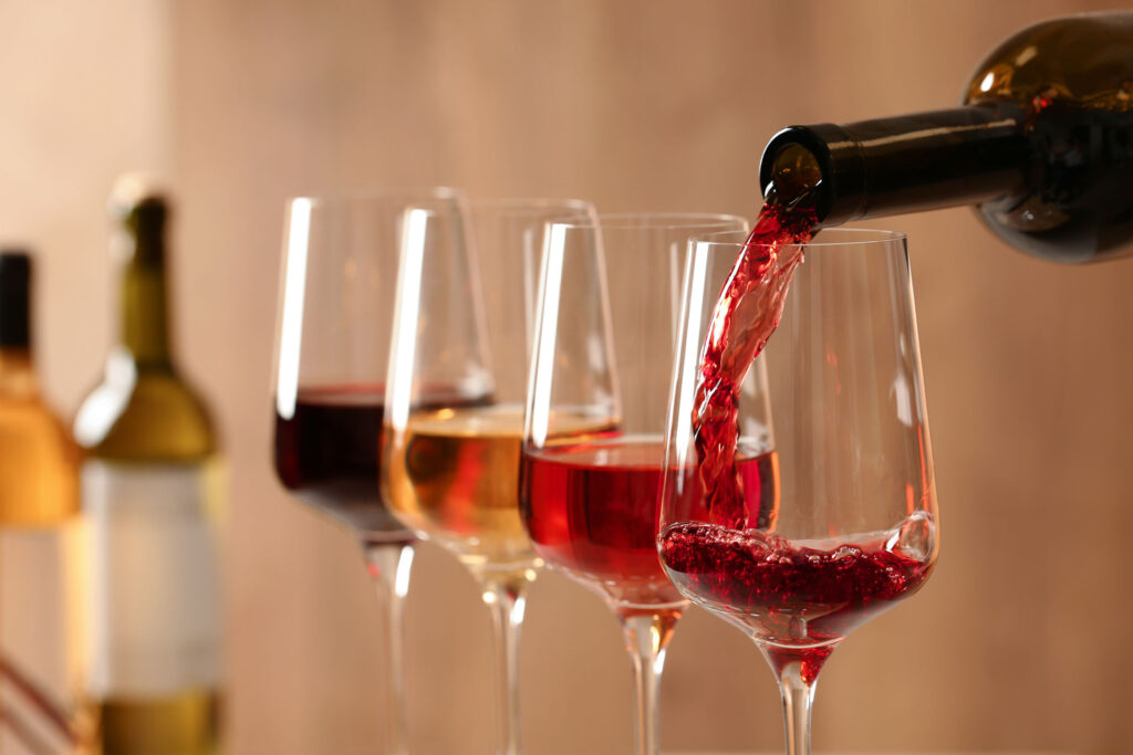 Red wine being poured out of a bottle into a wine glass that already has a little wine in it. Next to the glass are three other glasses with red wine, white wine, and rose in them. In the blurred background are two bottles of wine.