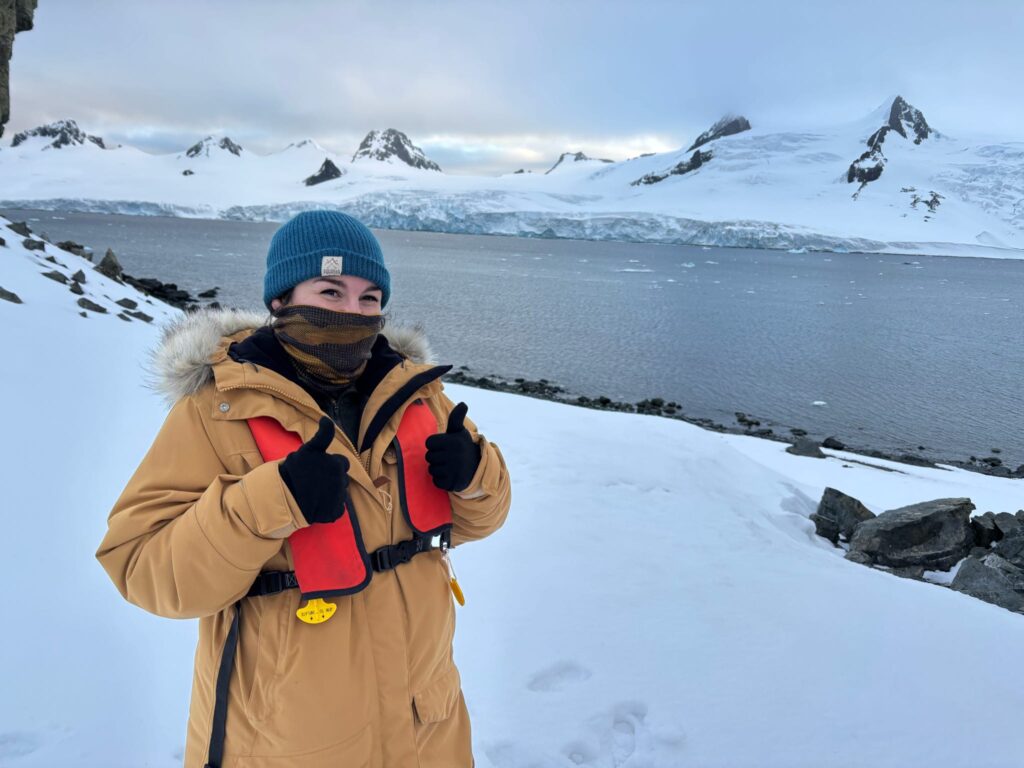 A person wearing lots of winter clothing and a mask gives a double thumbs up sign on a snow-covered landscape with water and mountains in the background.