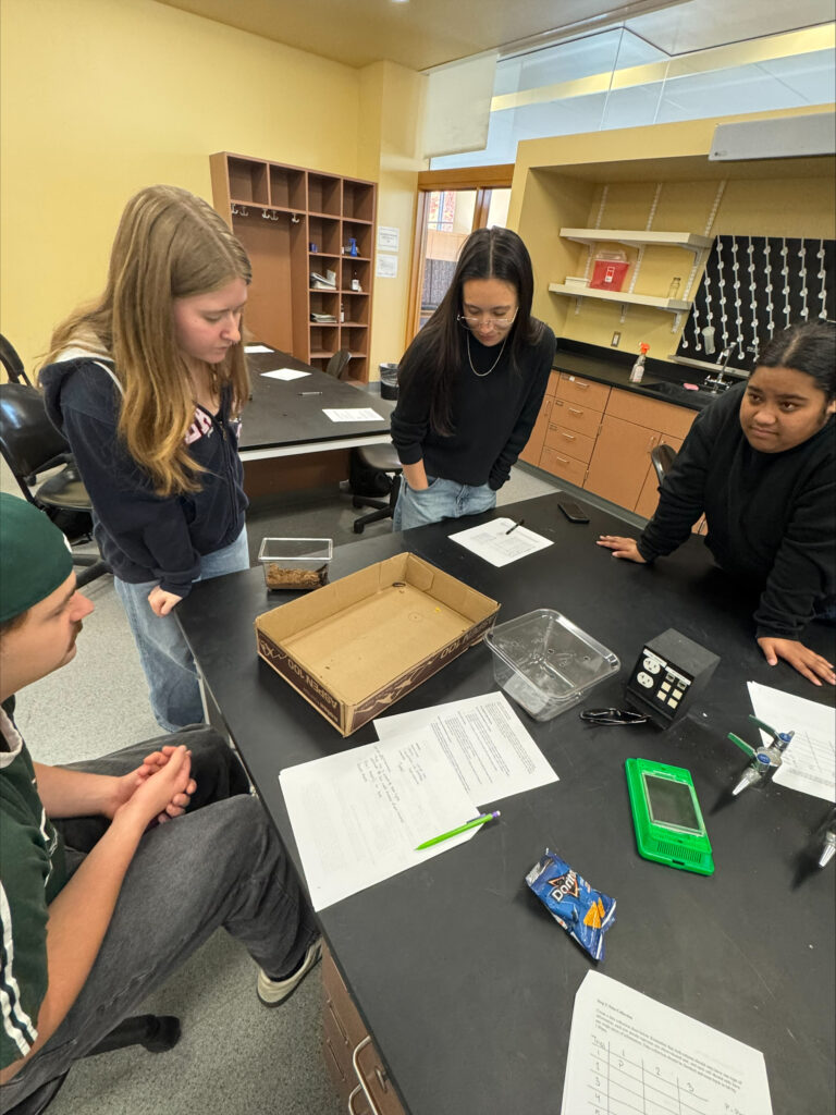 Four people gather around a table in a lab setting. On the table in front of them are papers, a cardboard box, a bag of Doritos chips, and plastic containers.