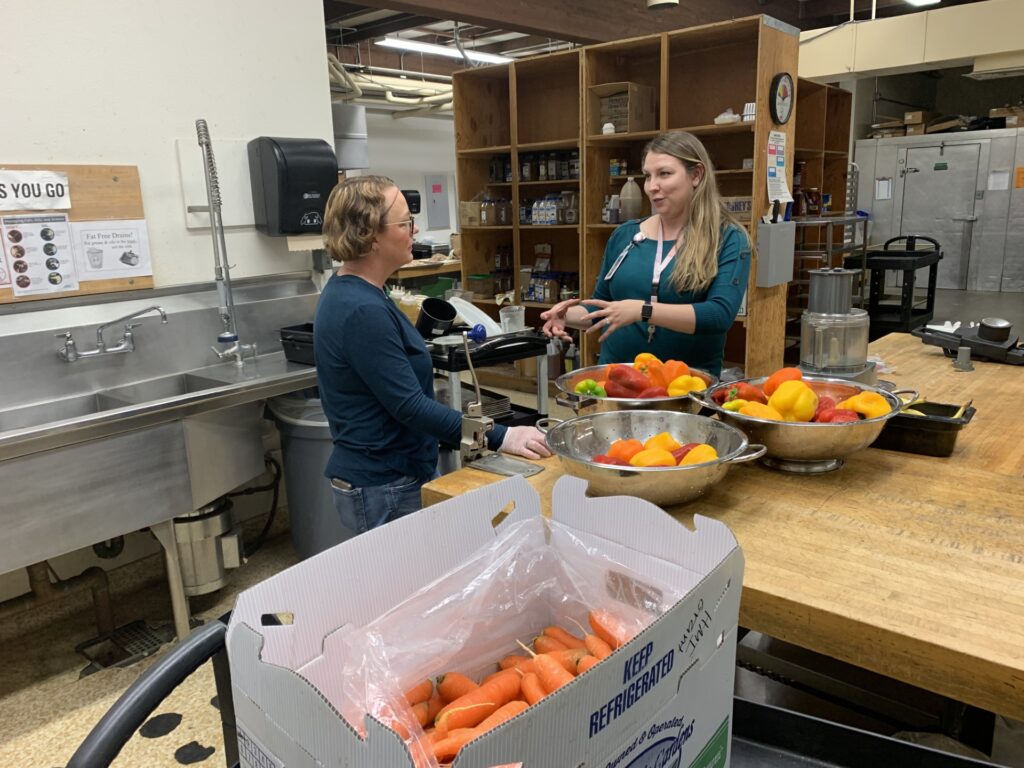 Two people talk in a food processing kitchen. Mixed color peppers line the butcher-block countertop. A full box of carrots is open.
