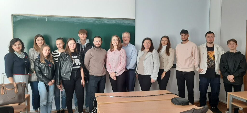 A large group of about 14 people stands behind a couple of tables and in front of a green chalk board.