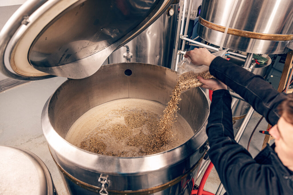 A person pours a grain from a bag into a large metal vat.
